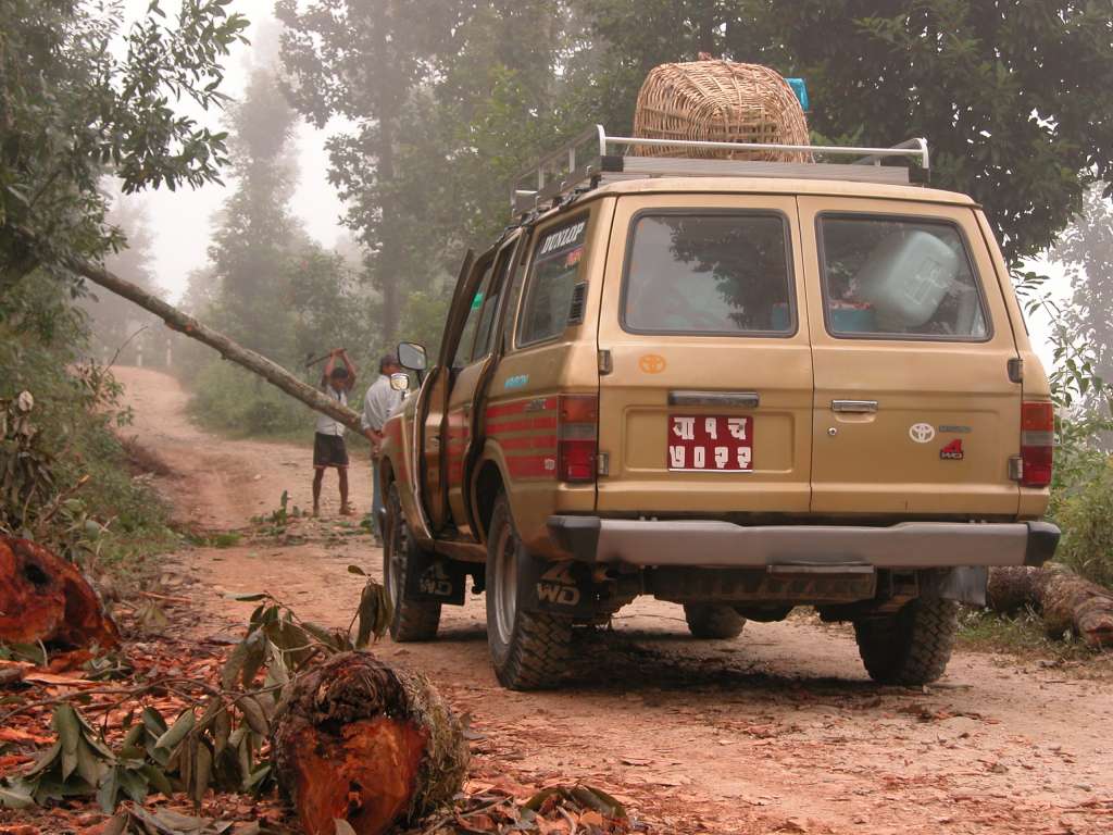 Manaslu 01 01 Landcruiser Stopped By A Tree On The Road We left Kathmandu in the dark at 5:15 and drove past people walking, bicycling and even jogging. At 6am we crested the lip of the valley and left Kathmandu behind. We continued west along the paved Prithvi Highway passing the ubiquitous Tata trucks and buses. We left the highway at 7:30 and drove 17km on a paved road to Dhading Besi, turned left and followed the unpaved road up through the early morning clouds. A small traffic jam occurred when a large tree fell across the road and had to be cut by one of the local young men.